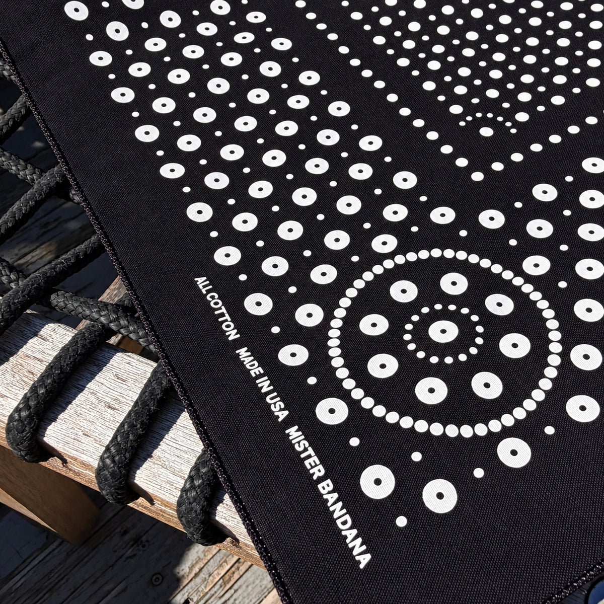 Rivet bandana in Black with white dot print, corner detail showing the All Cotton / Made in USA / Mister Bandana label, on a weathered wood bench