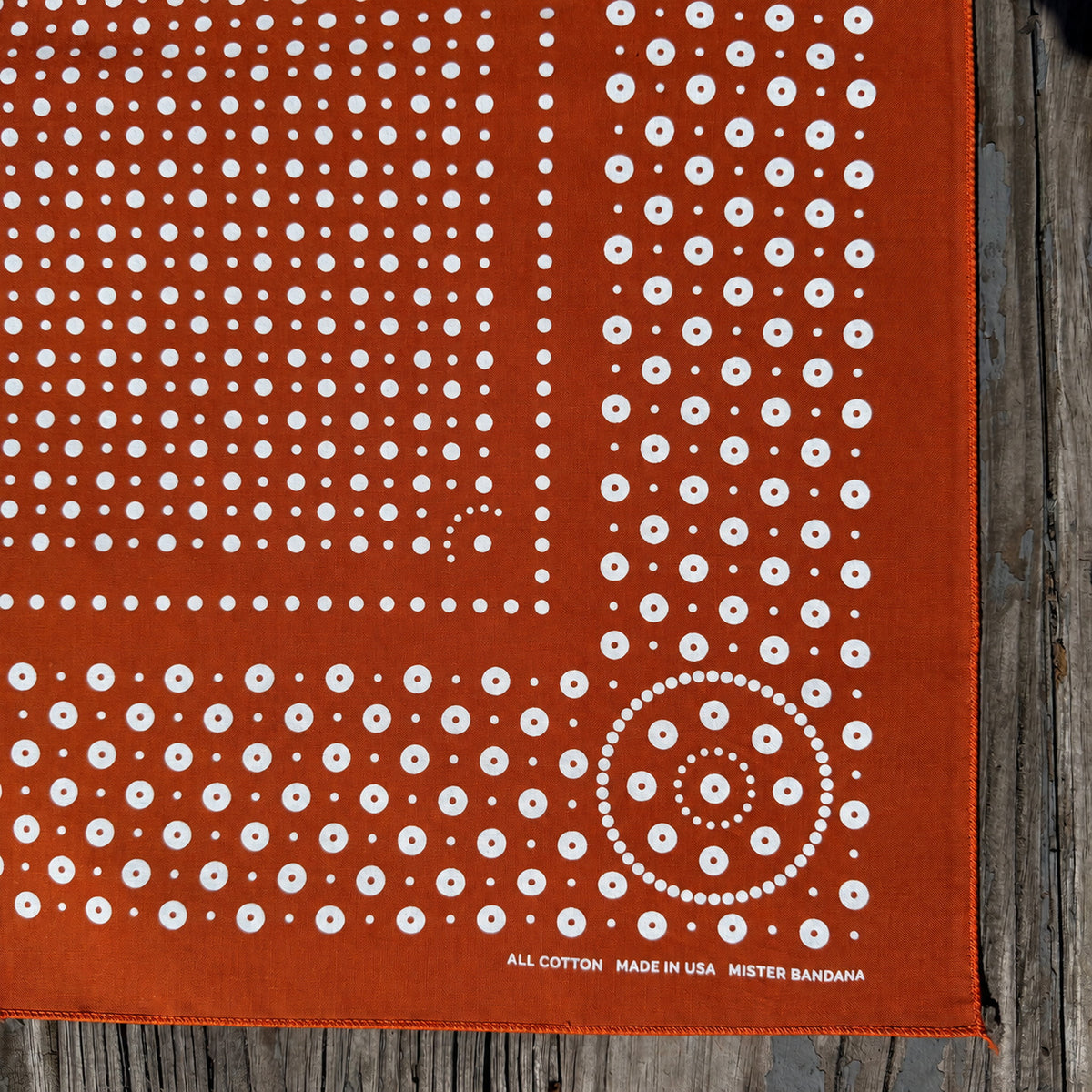 Rivet bandana in Burnt Orange with white dot print, corner detail with stitched hem, laid flat on weathered wood